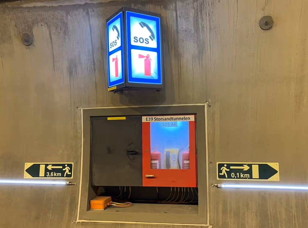 Nødtelefonskilt og røykdykkingsutstyr i E39 Storsandtunnelen, med skilt som viser nødutganger 0,1 km til høyre og 3,6 km til venstre.