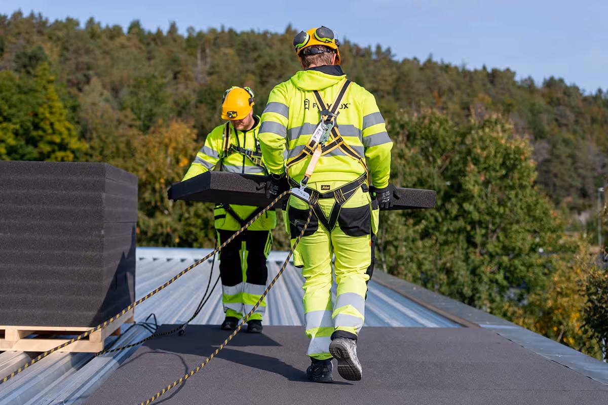 To byggearbeidere i høy synlighet klær og hjelm bærer isolasjonsplater på et tak under utendørs arbeid.
