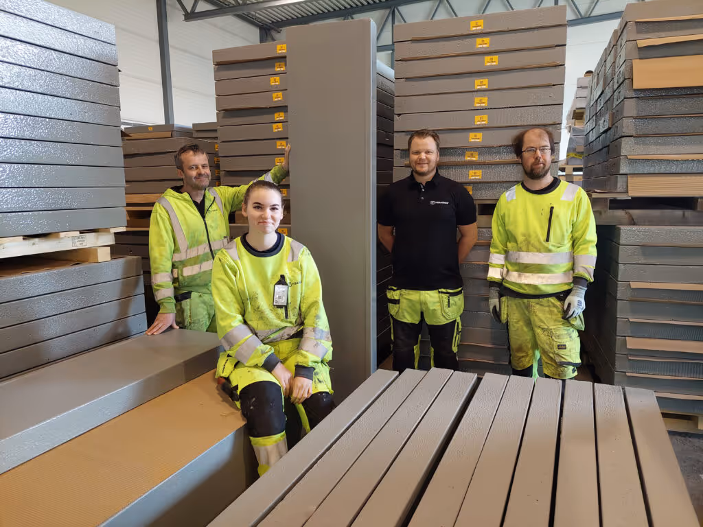 Fire fire-fire workers in yellow arbeidsklær som poserer foran stabler av store grå isolasjonspaneler.