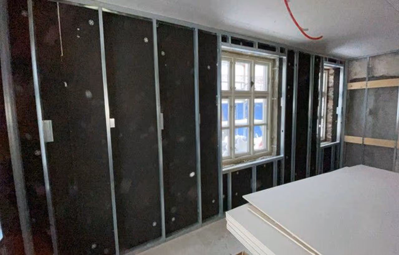 Innvendig byggearbeid med metalstendere montert på veggene og en stabel med gipsplater i et rom, med vinduer i bakgrunnen.