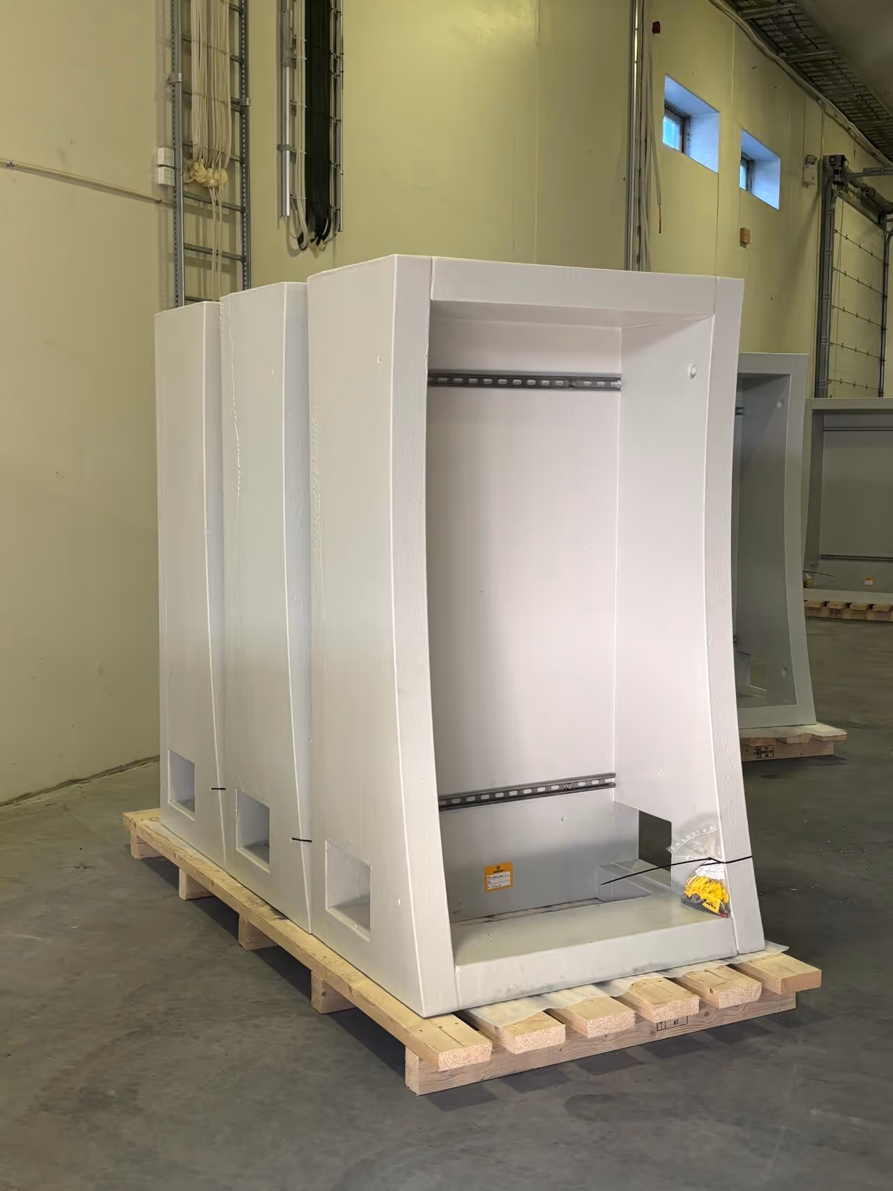 Tre store hvite skap med åpne fronter stående på en trepalle i et lagerrom med grå betonggulv og gulmalte vegger.