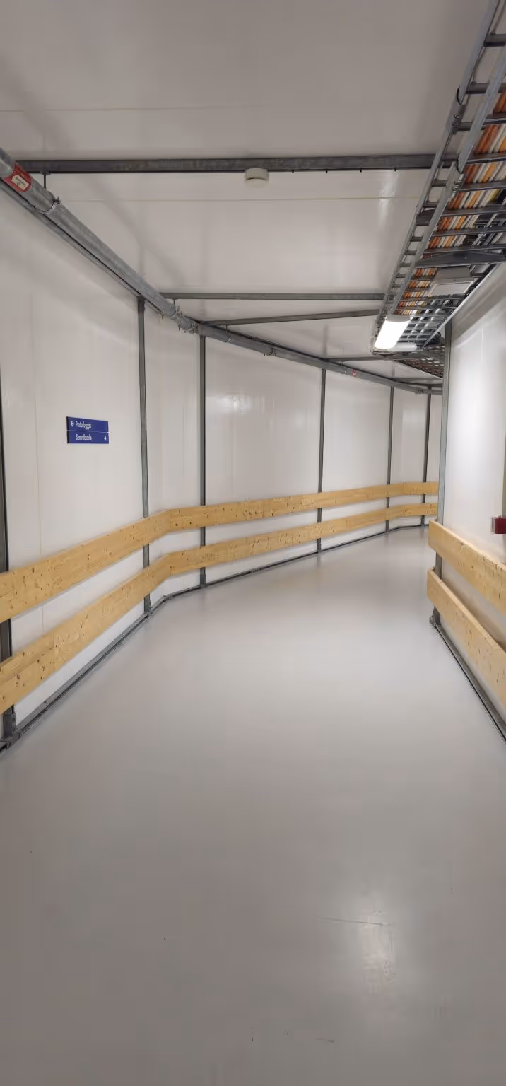 Innendørs korridor med hvite vegger, gulv og tak, tregelendere og en blå skilt på veggen.