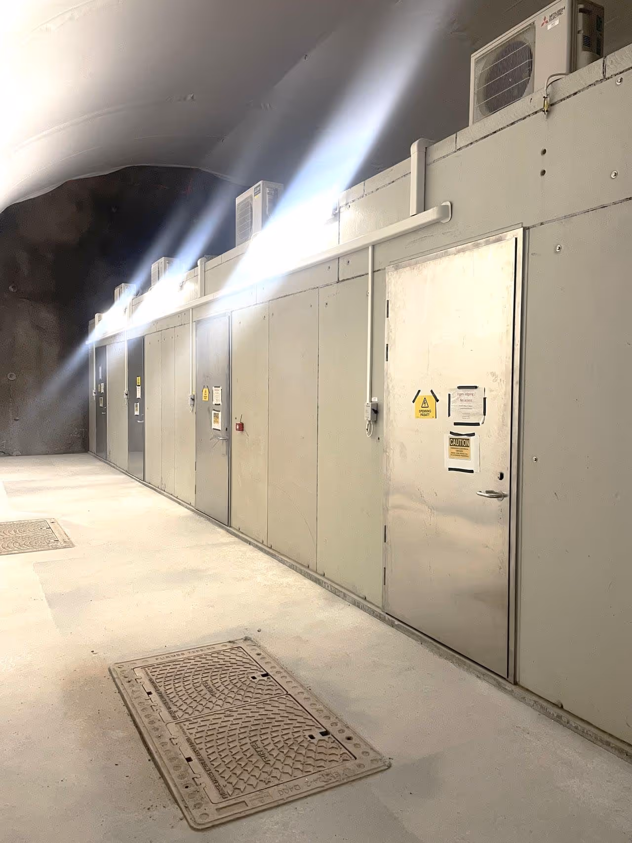 Langt smalt rom med metallsdører og luftkondisjoneringsenheter på toppen, med skilt som advarer om fare.