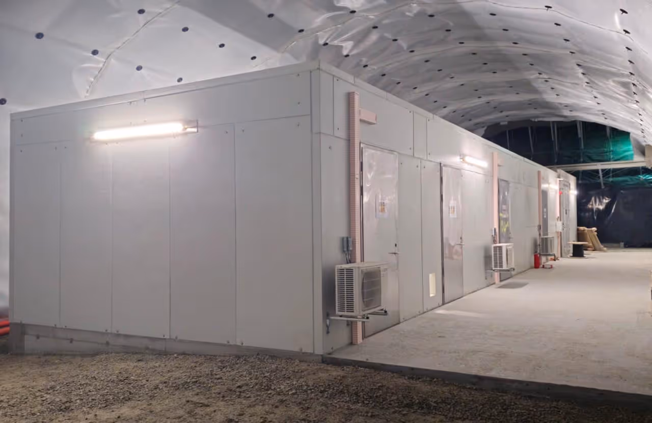 Teknisk bygg med flere dører og klimaanlegg under et hvitt, buet tak i et industrielt miljø.