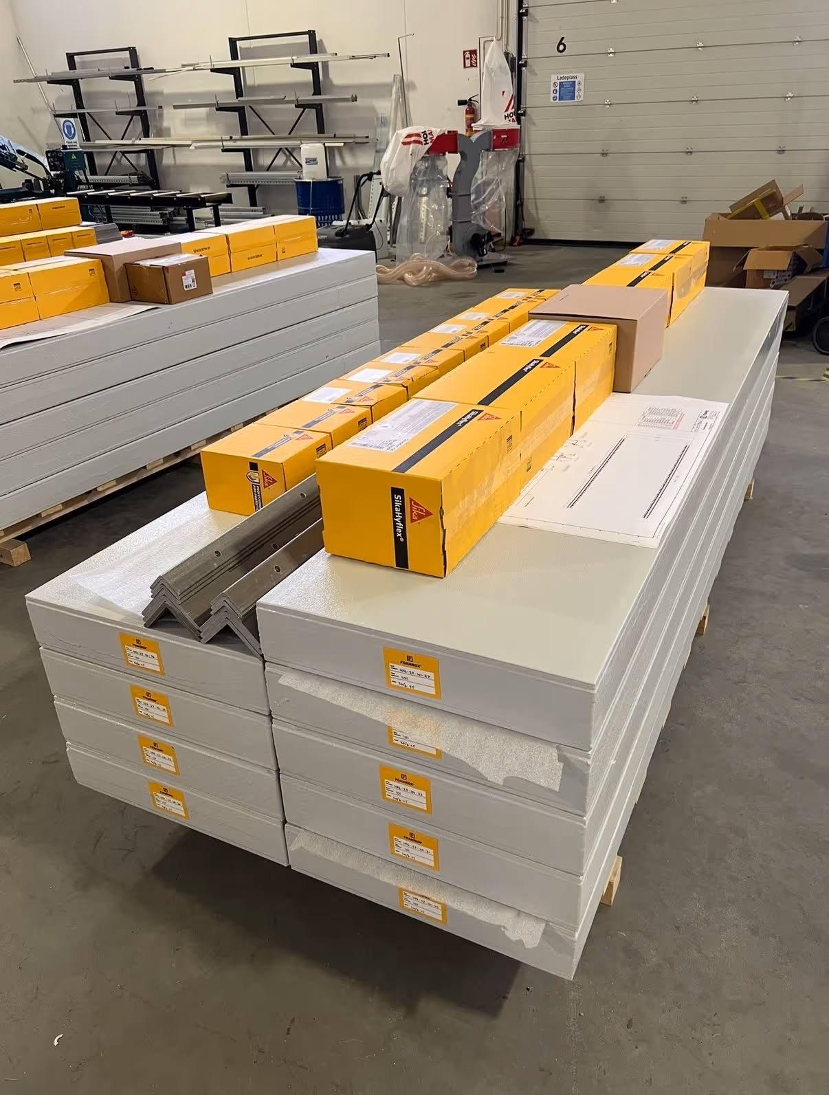 Stabler med hvite byggeplater og gule esker med verktøy plassert oppå i et lagerrom.