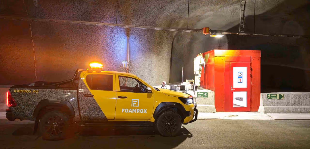 Foamrox-gul og svart pickup med blinkende oransje taklys parkert i en underjordisk tunnel ved siden av en nød-kiosk i rødt, med en person i verneutstyr som står ved kiosken.