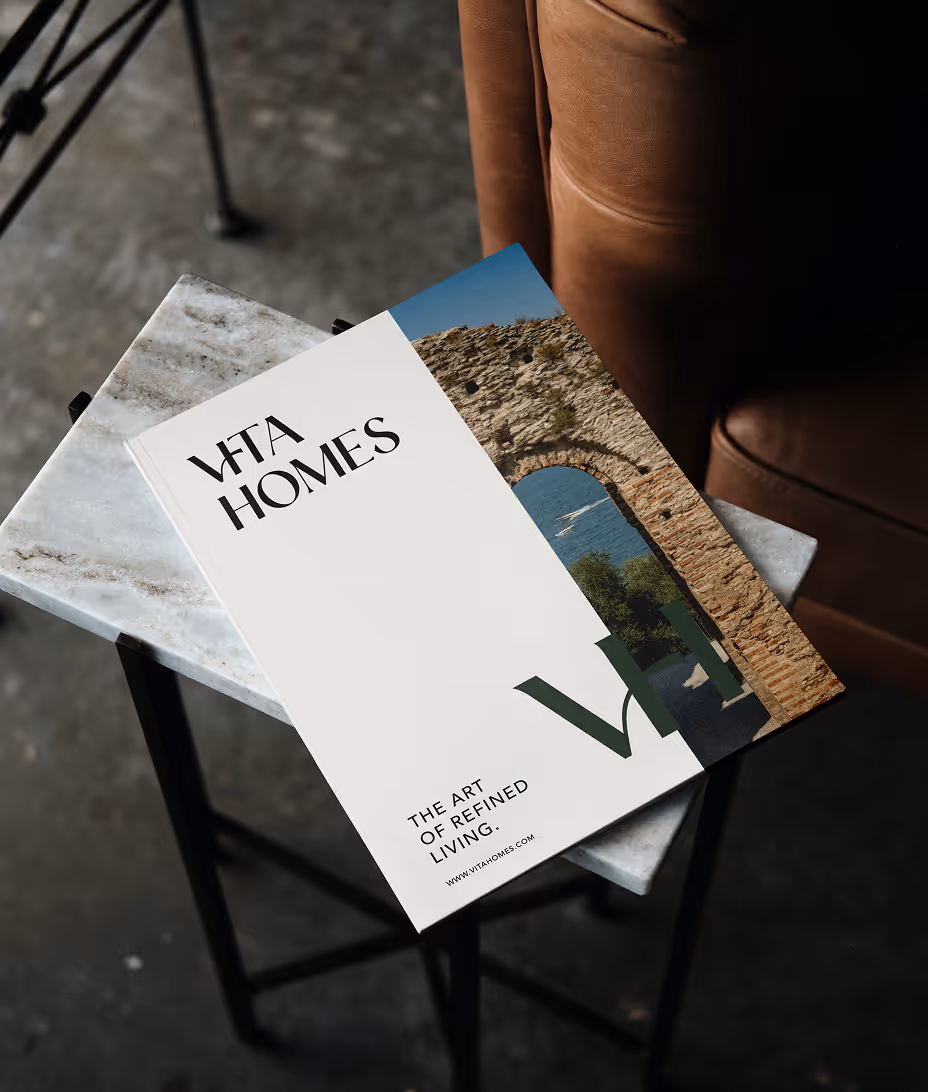 Vita Homes brochure on a marble table beside a brown leather chair, featuring a stone arch photo.