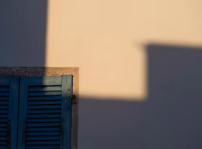 Blue shuttered window partially covered by shadow on a beige wall in soft light.