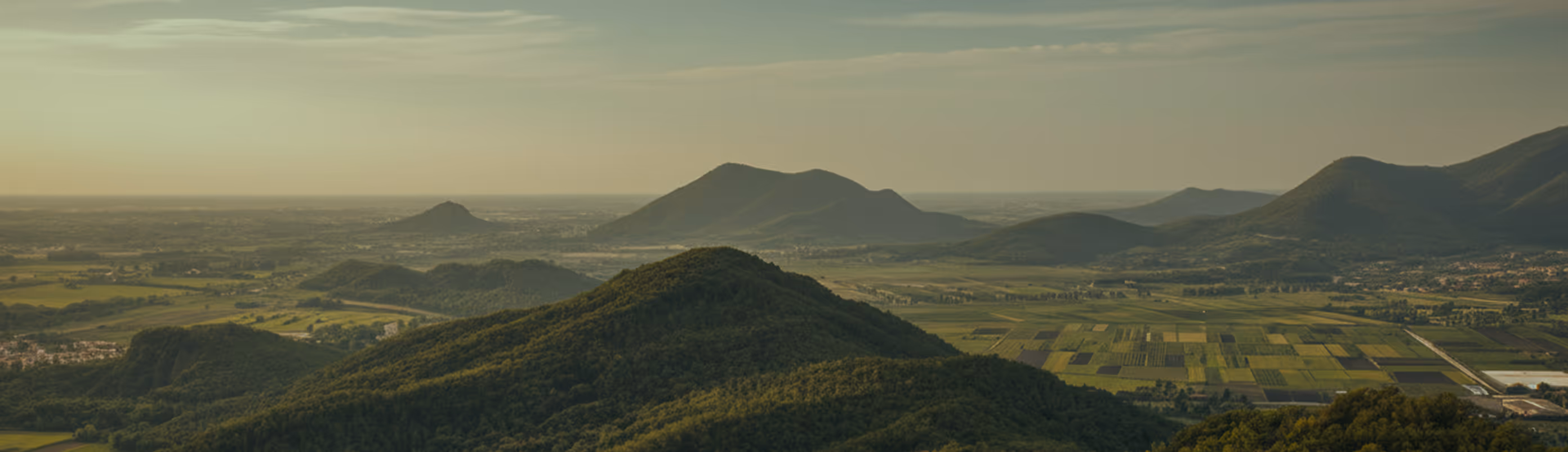 Panoramic view of green hills with farmland patches in a valley under a cloudy sky.