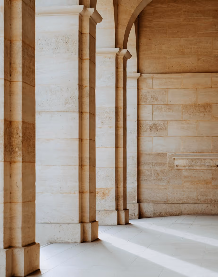 Stone pillars with arches casting long shadows on a tiled floor in a sunlit corridor.