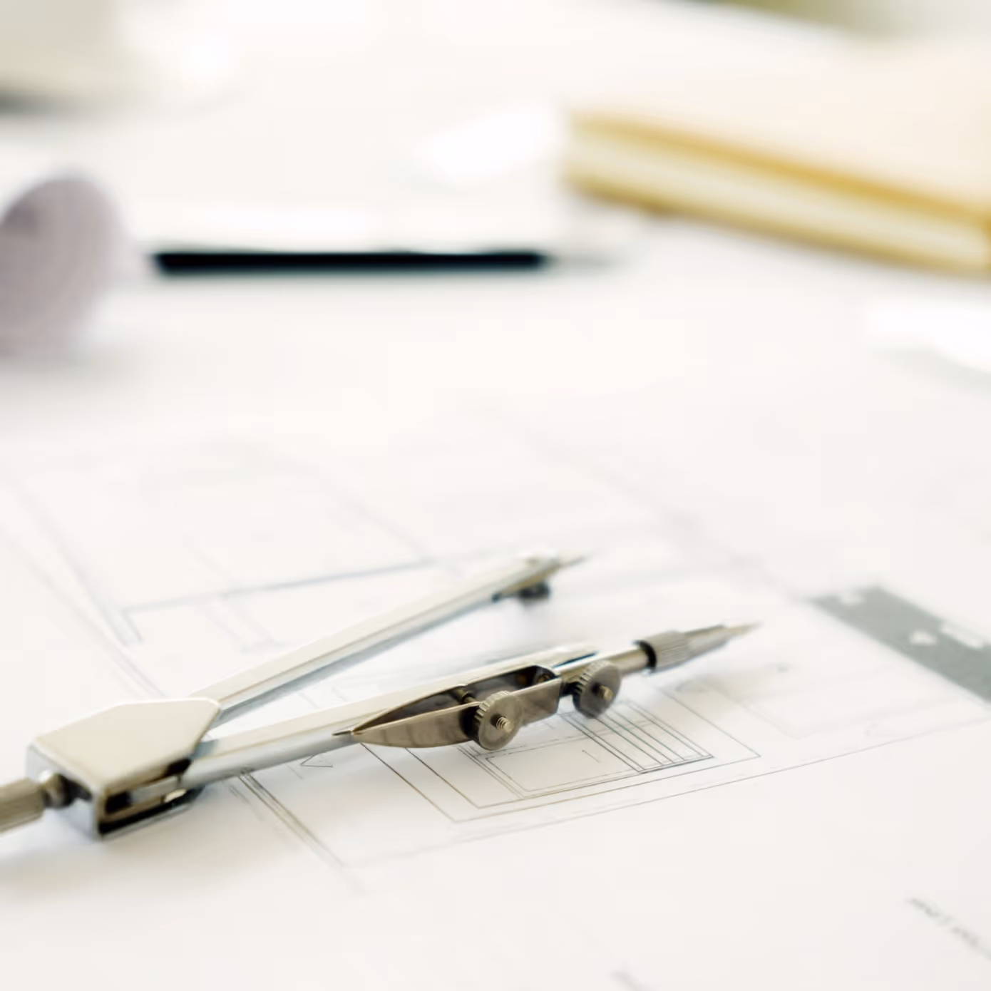 Metal compass resting on architectural blueprints with pen and notebook blurred in background.