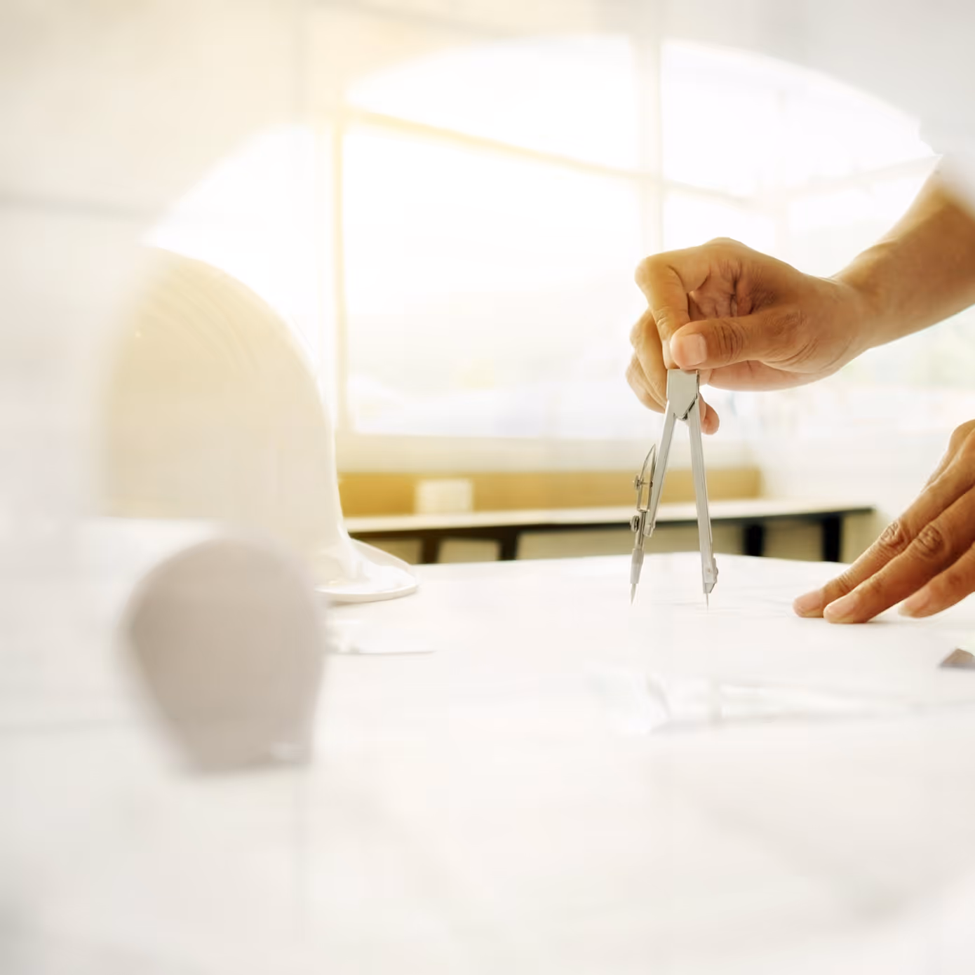 Hand using a compass on blueprints with a white hard hat blurred in the foreground.