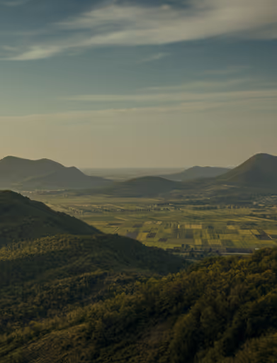 Wide valley with patchwork fields surrounded by forested hills under a partly cloudy sky.