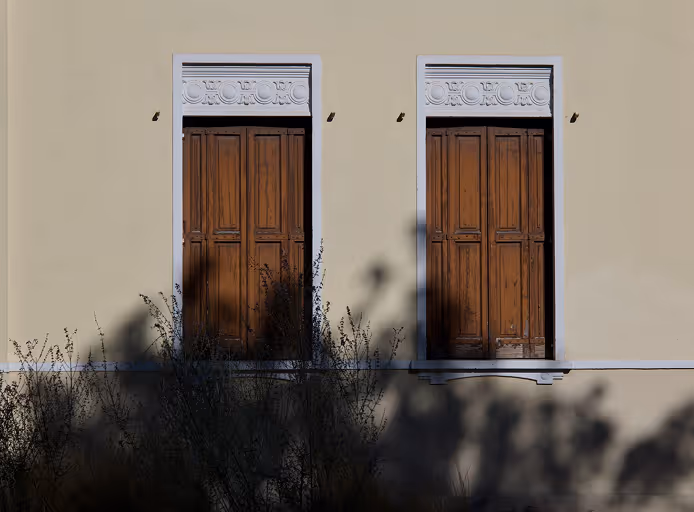 Two closed wooden windows with white decorative frames on a beige wall, shadows of plants below.