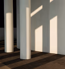 Sunlight casts geometric shadows from large columns onto a dark tiled floor and a white wall.