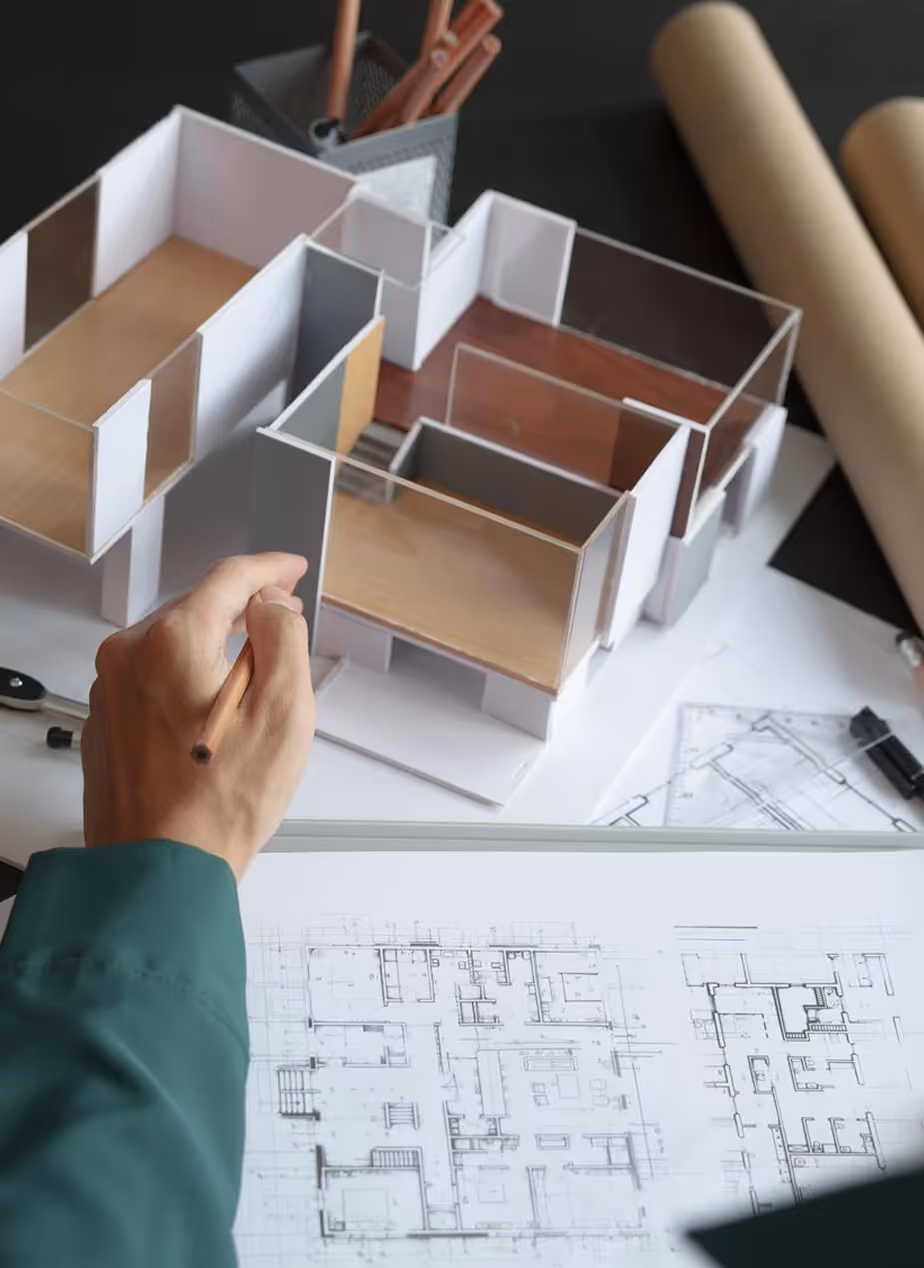 Person holding pencil near architectural model and floor plans on a desk with tools.