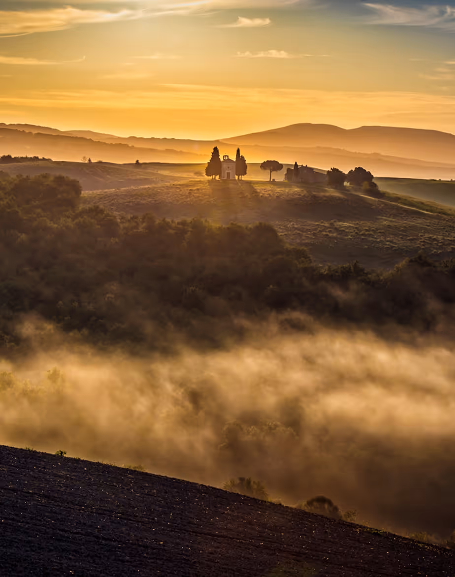 Sunrise over misty hills with a small chapel and trees on a ridge in warm golden light.