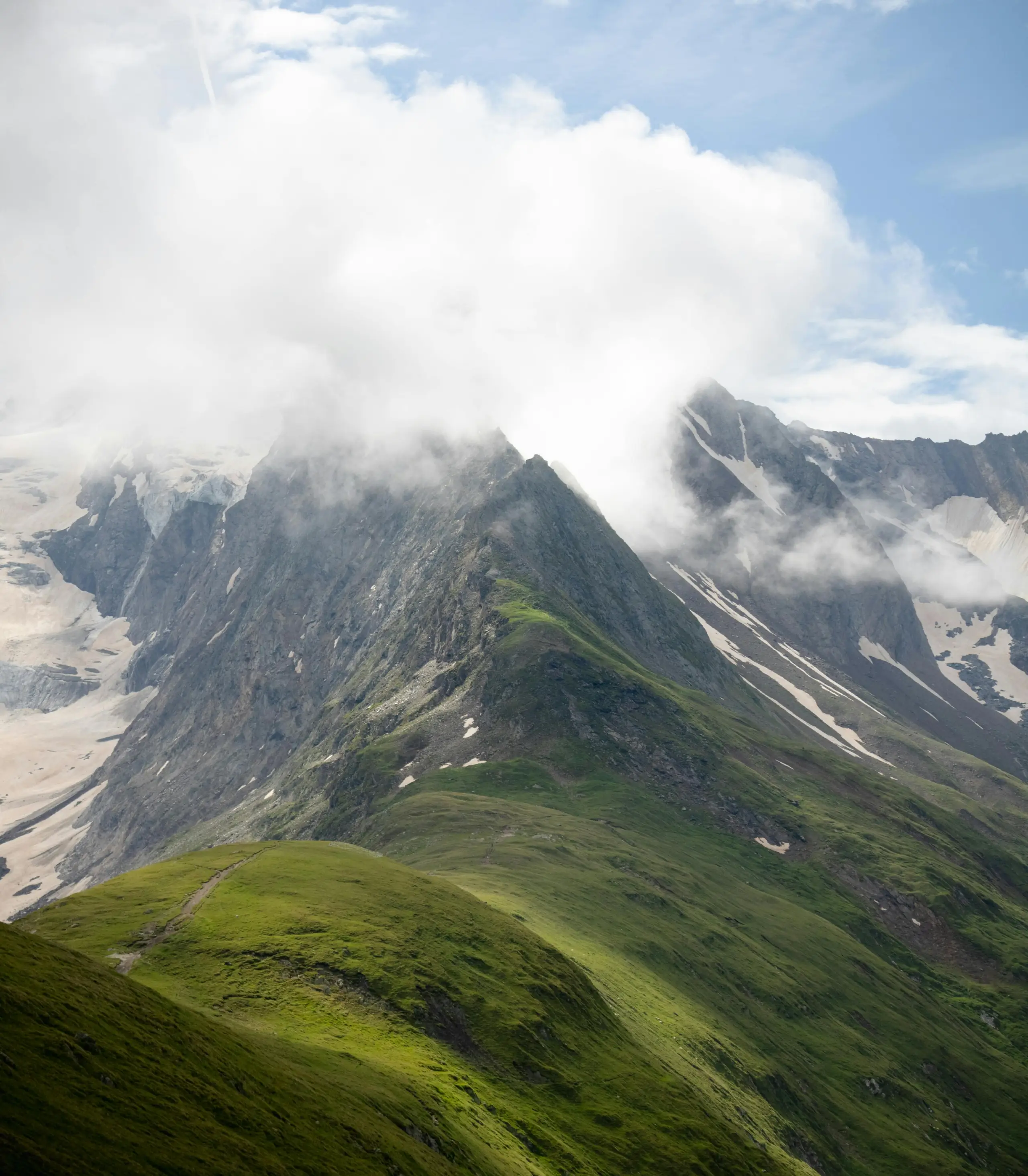 Mountain peaks with green slopes partially covered by mist and clouds under a blue sky.