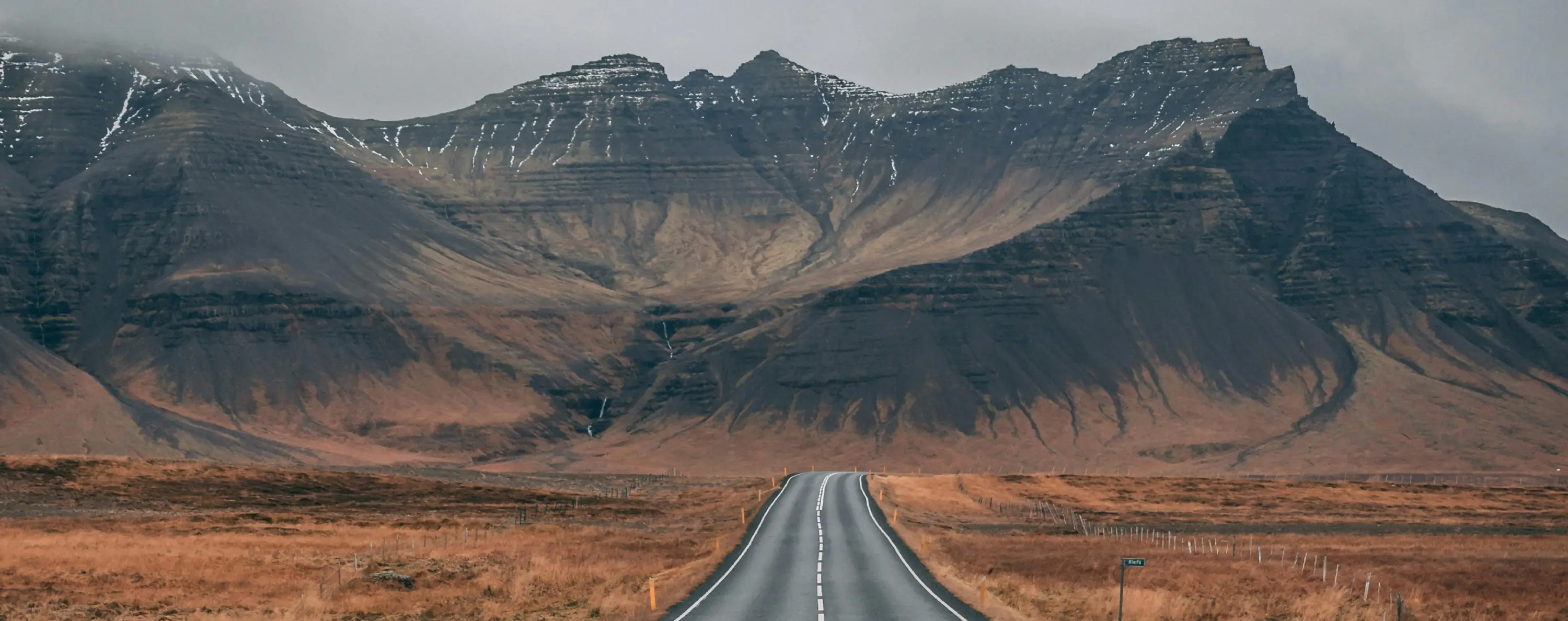 Empty road leading towards large rocky mountains with patches of snow under a cloudy sky.