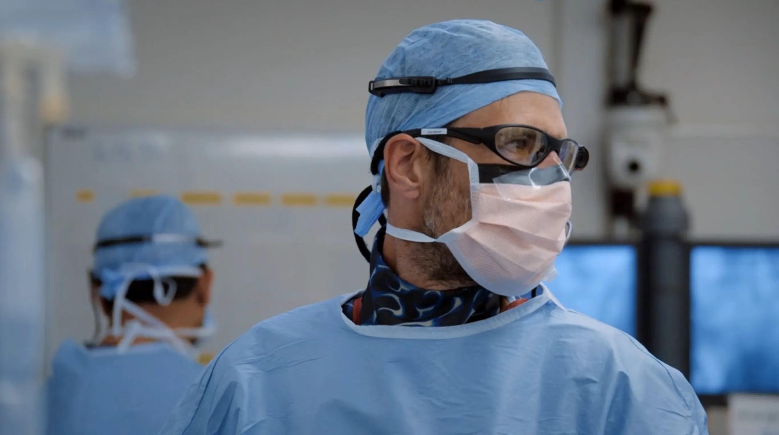 Chris Mitchell wearing blue surgical scrubs, mask, protective glasses, and cap in an operating room.