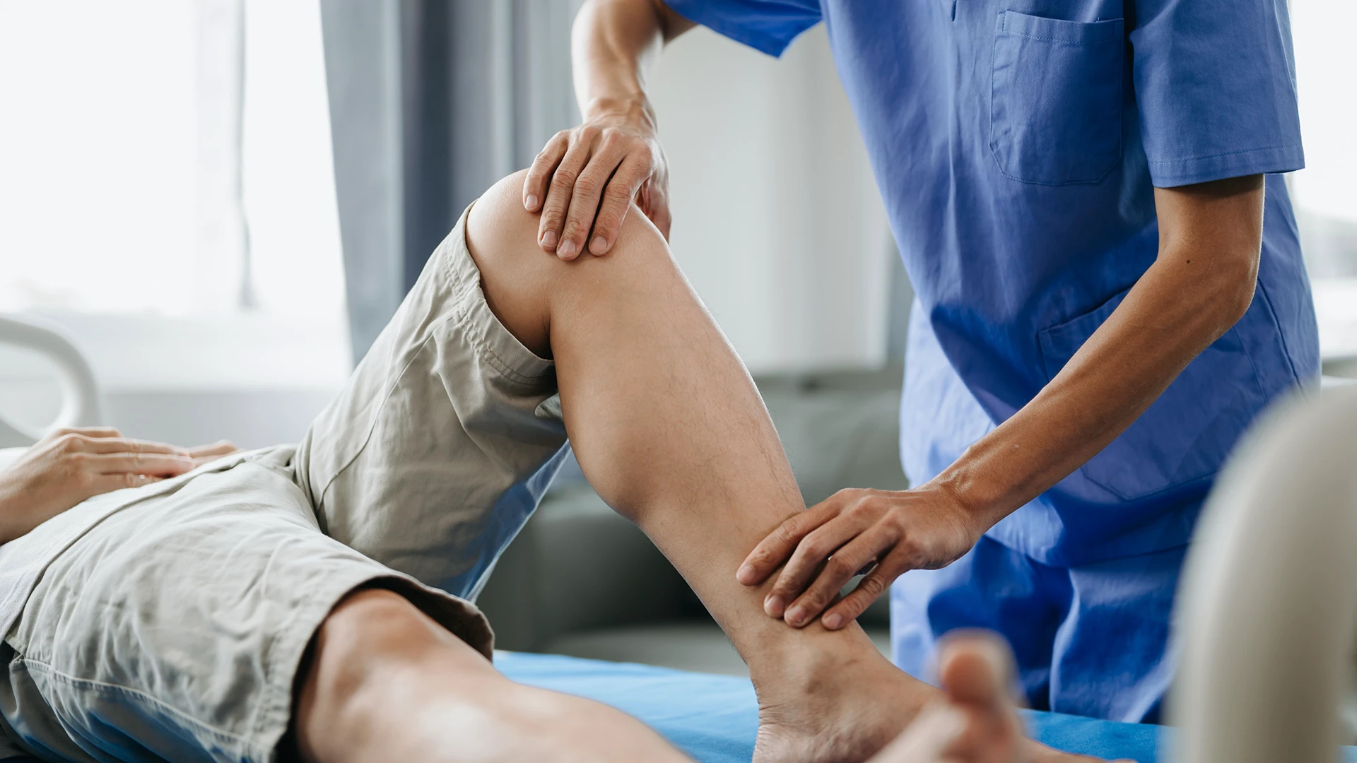 Physical therapist in blue scrubs examining a patient's bent knee on a treatment table.