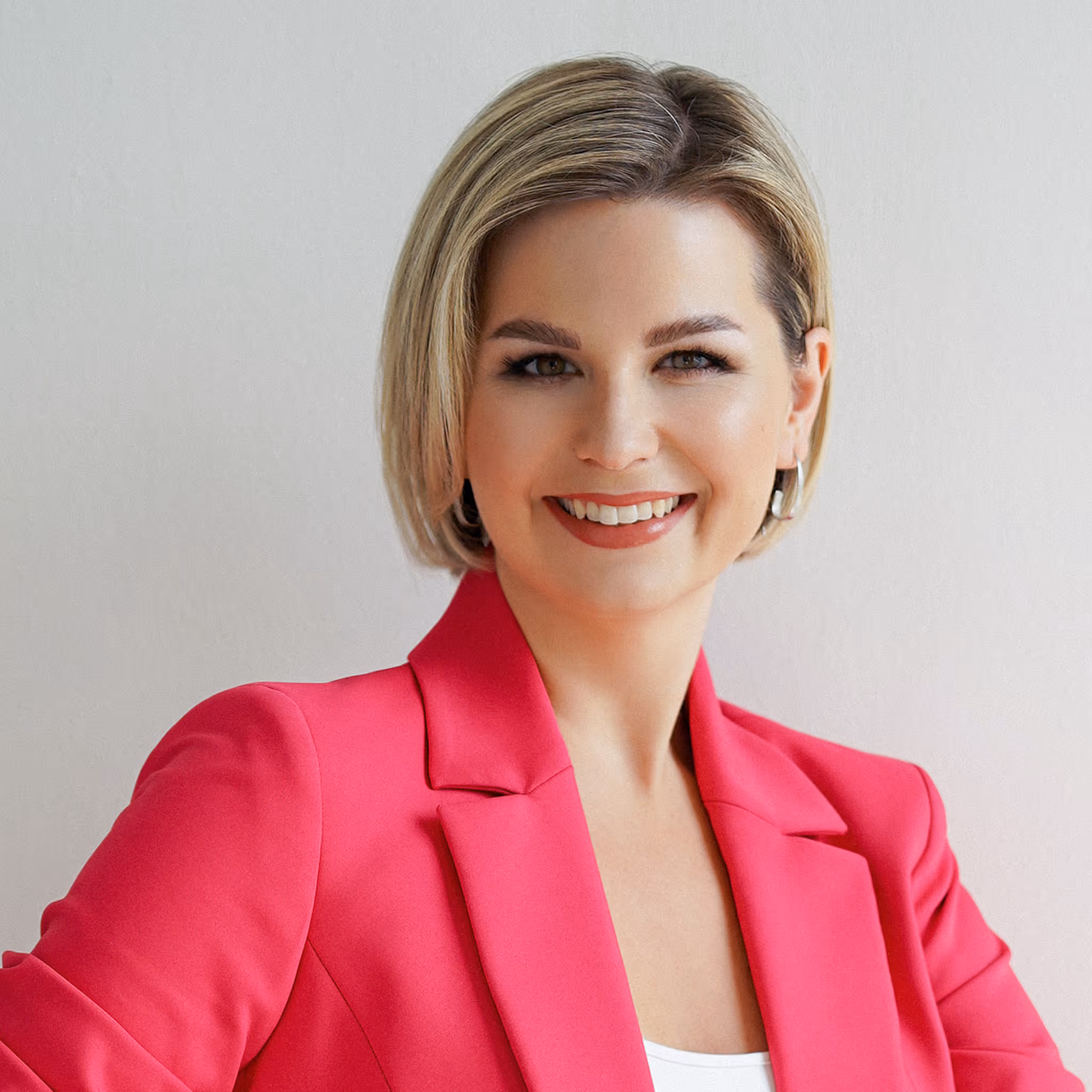 Lächelnde Frau mit kurzem blondem Haar, die ein pinkfarbenes Sakko und silberne Ohrringe trägt.