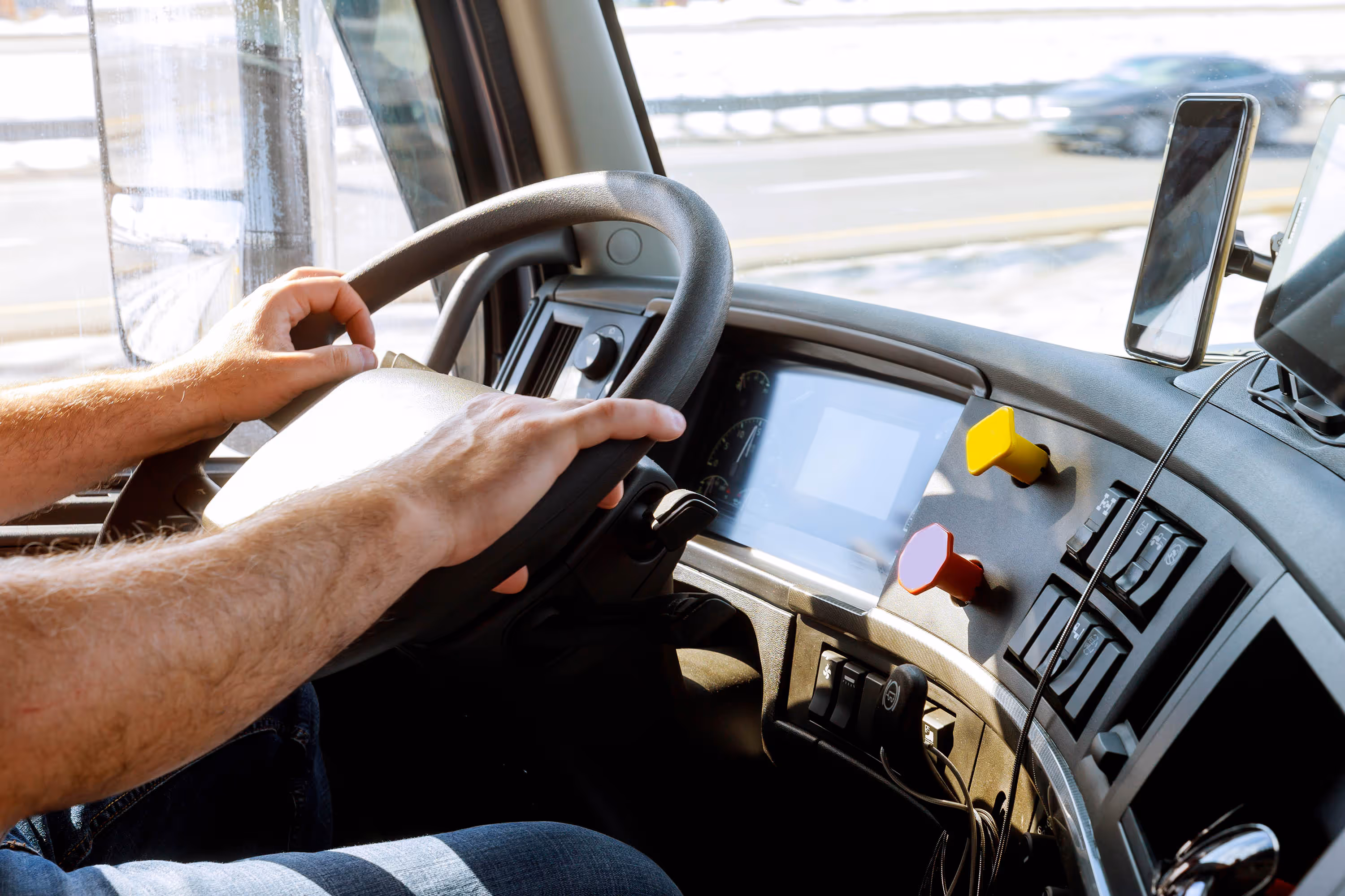 Hands of a driver gripping a truck steering wheel with dashboard controls and a mounted smartphone in view.
