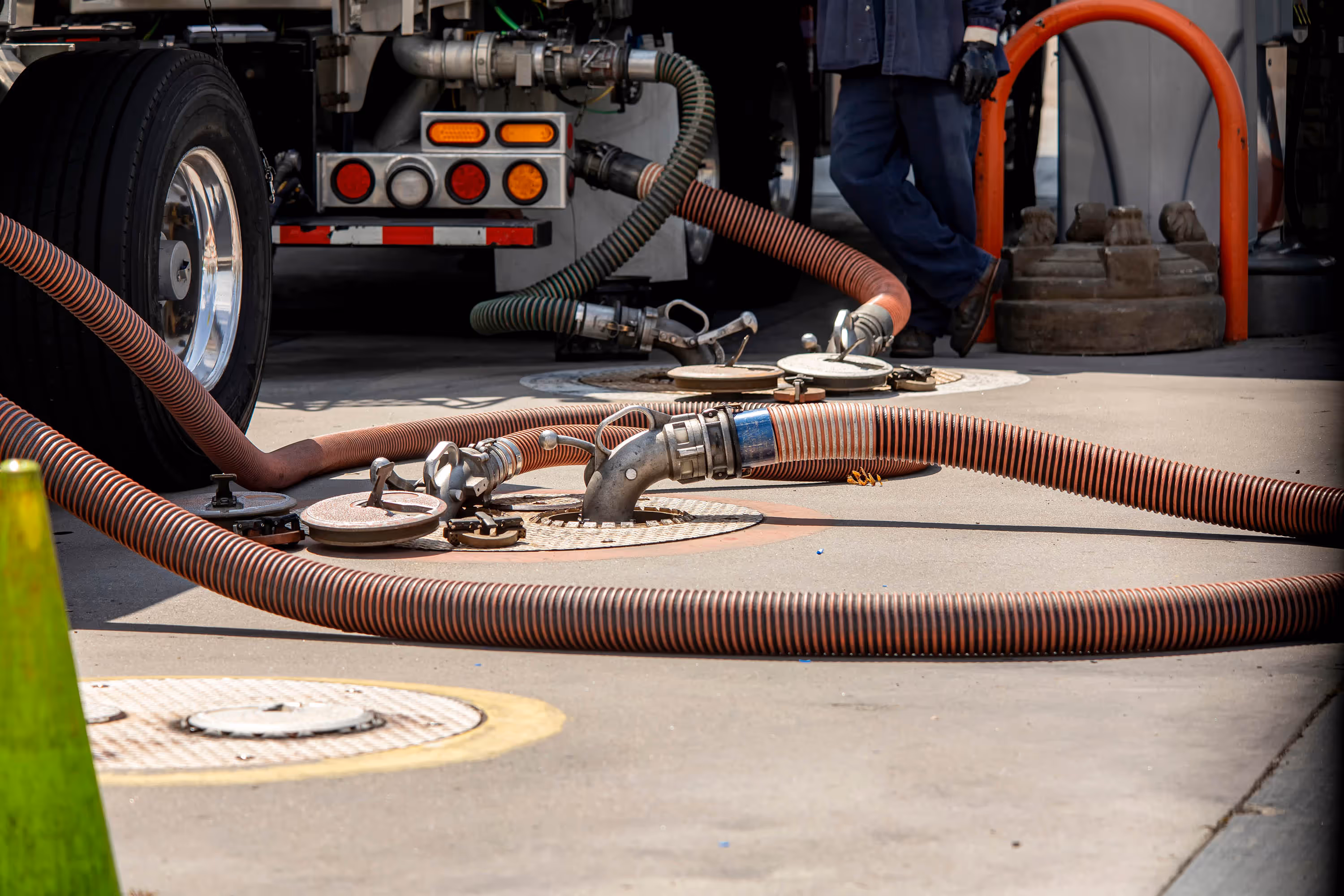 Fuel truck hoses connected to ground-level intake valves with a worker standing nearby.