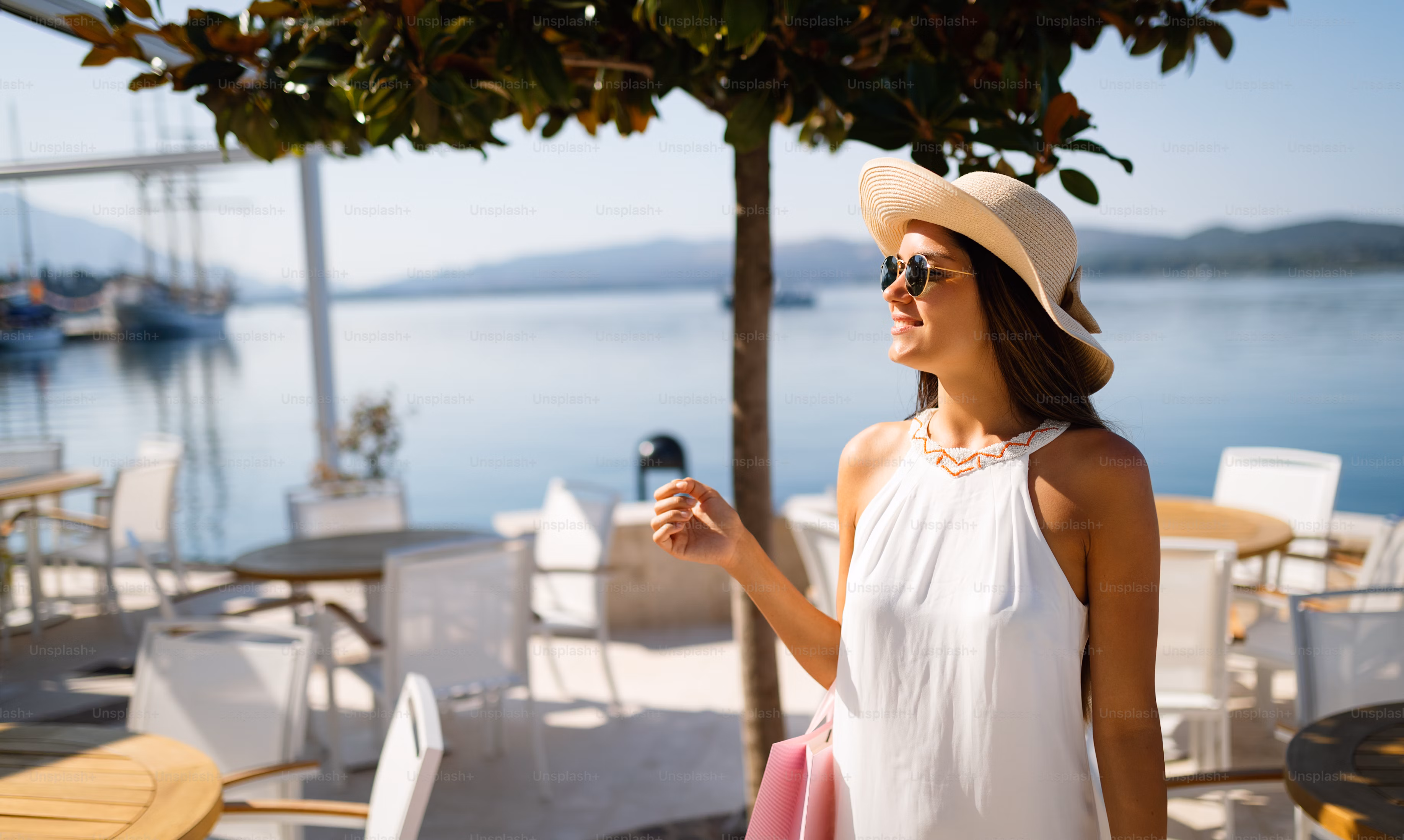 Woman in a white dress and sunhat smiling outdoors at a seaside cafe with tables, chairs, and a tree.