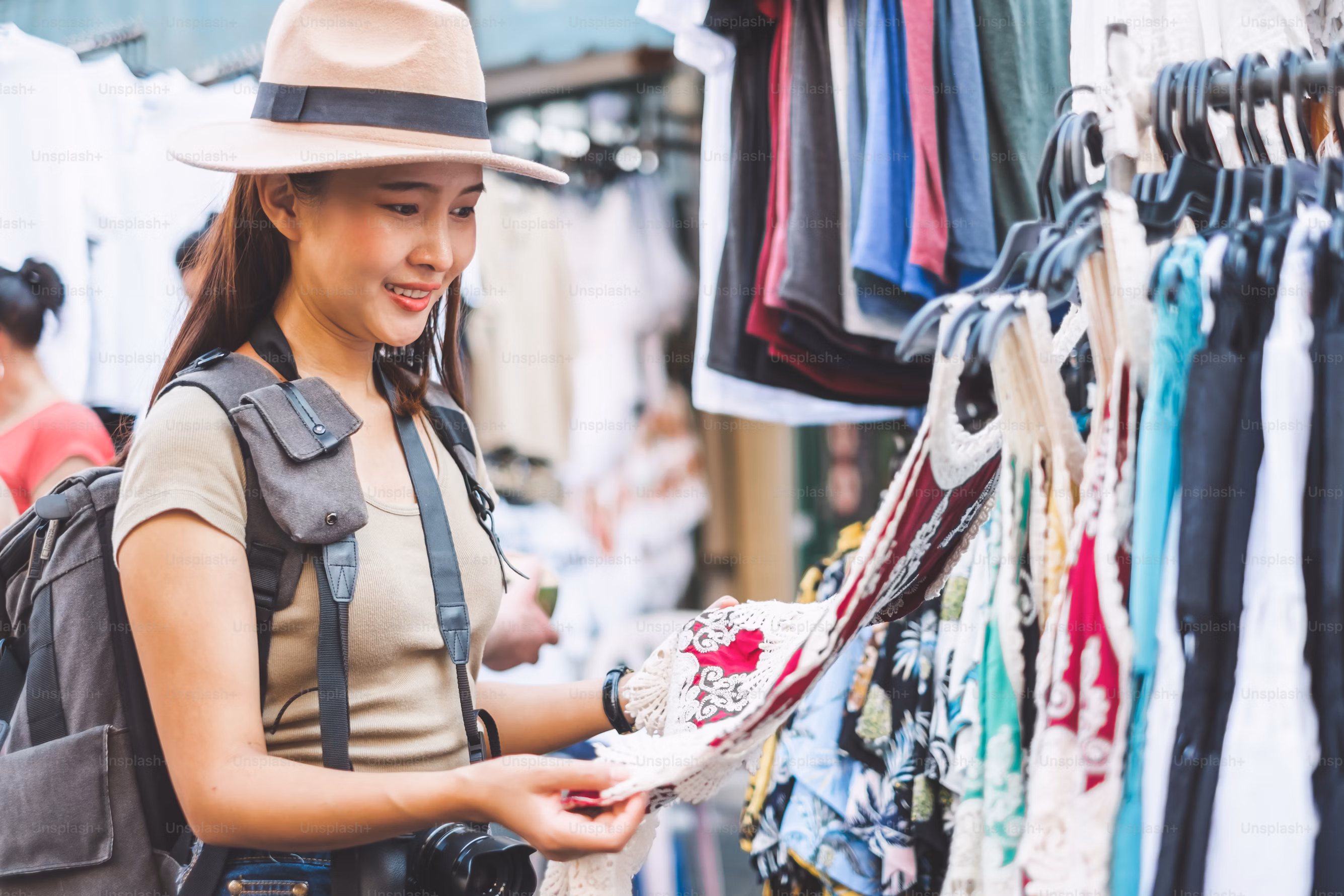 Smiling woman wearing a hat and backpack browsing colorful clothes on hangers at an outdoor market.