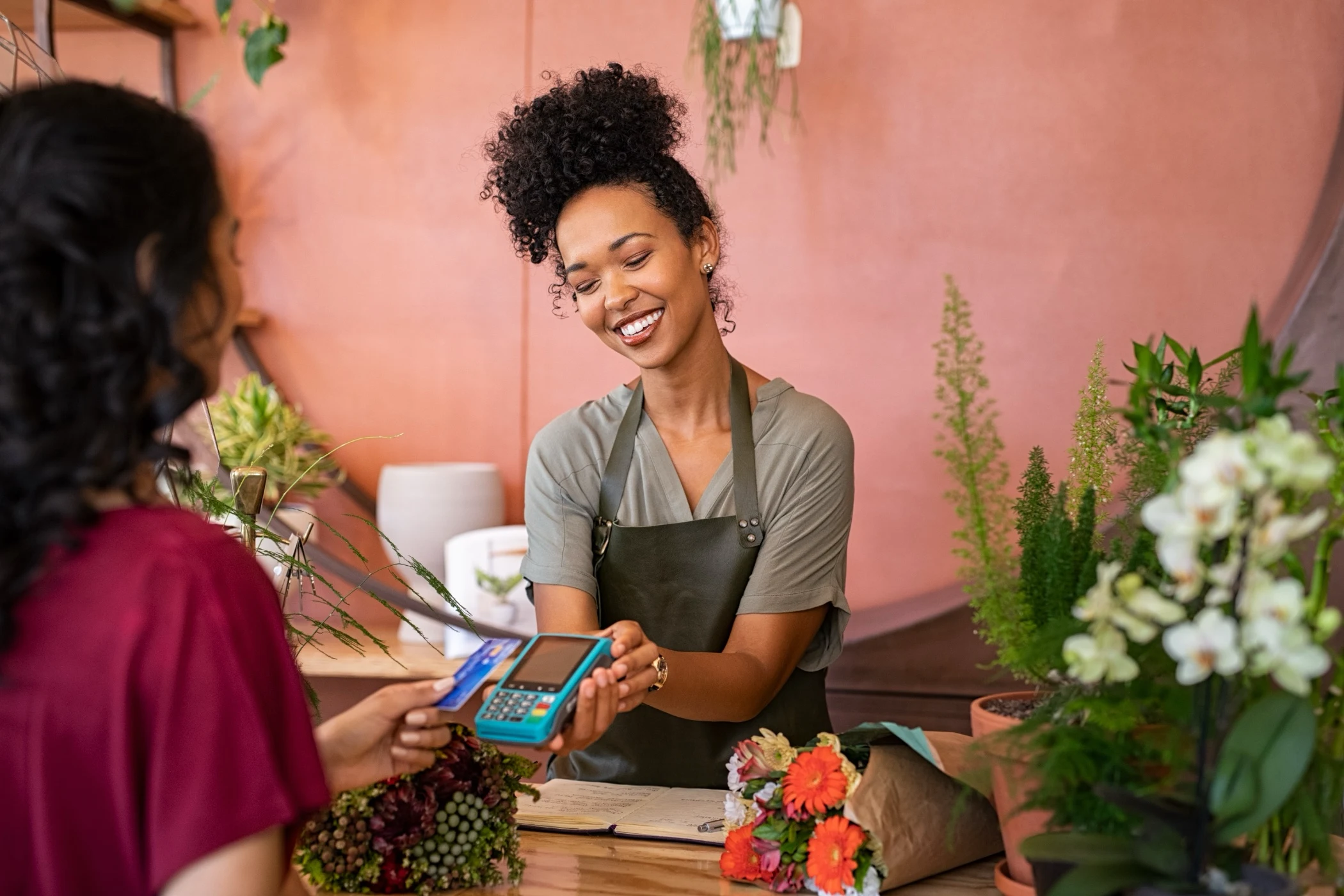 Smiling florist in an apron holding a card payment terminal while a customer holds a credit card over it in a flower shop.