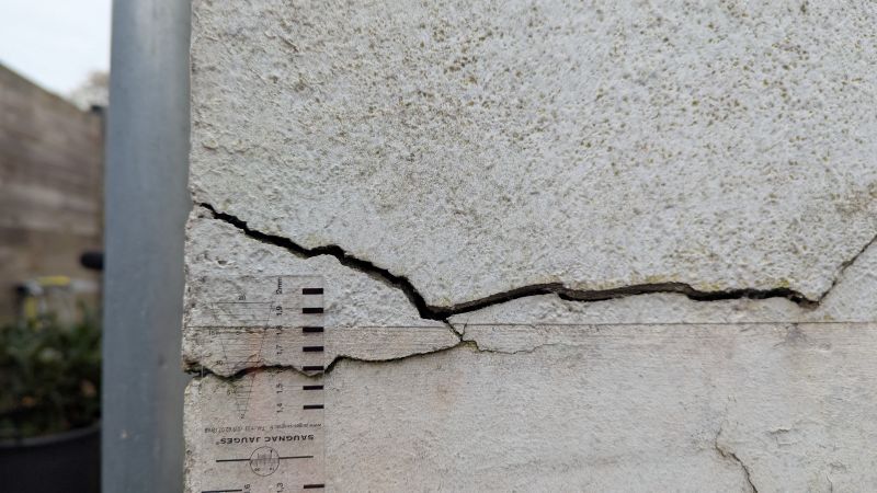 Gros plan sur un mur en béton fissuré avec une échelle de mesure placée verticalement à côté des fissures.