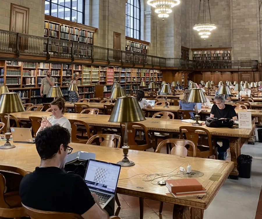 People studying and using laptops at wooden tables with brass lamps inside a large, classic library room filled with tall bookshelves.