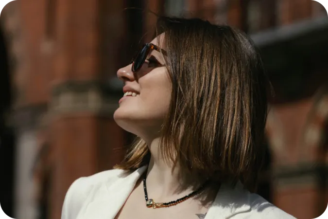 Woman with short brown hair wearing sunglasses and a white blazer, smiling and looking up outdoors.