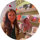 Smiling woman with long curly hair holding a book in a bookstore with colorful books on wooden shelves behind her.