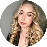 Portrait of a young woman with long curly blonde hair wearing a black top and a necklace, against a plain gray background.