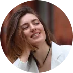 Smiling woman with shoulder-length brown hair wearing a white blazer and black necklace, outdoors.