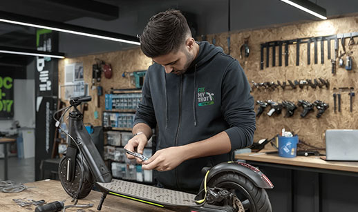 Un réparateur répare une trottinette électrique dans un atelier avec des outils accrochés au mur.