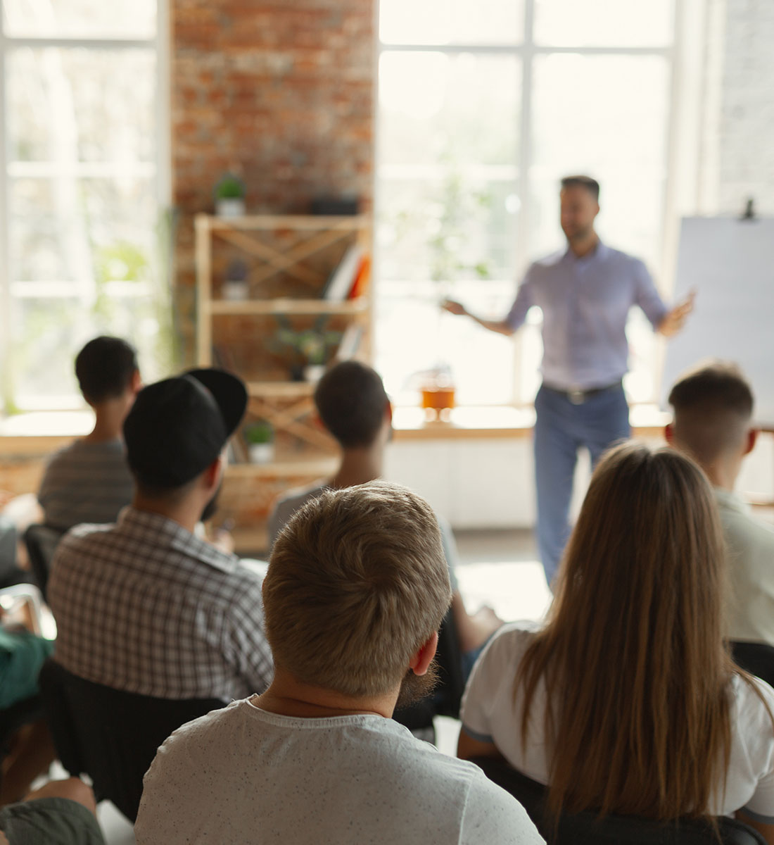 Un groupe de personnes assises regardant un homme donnant une présentation devant une fenêtre lumineuse.
