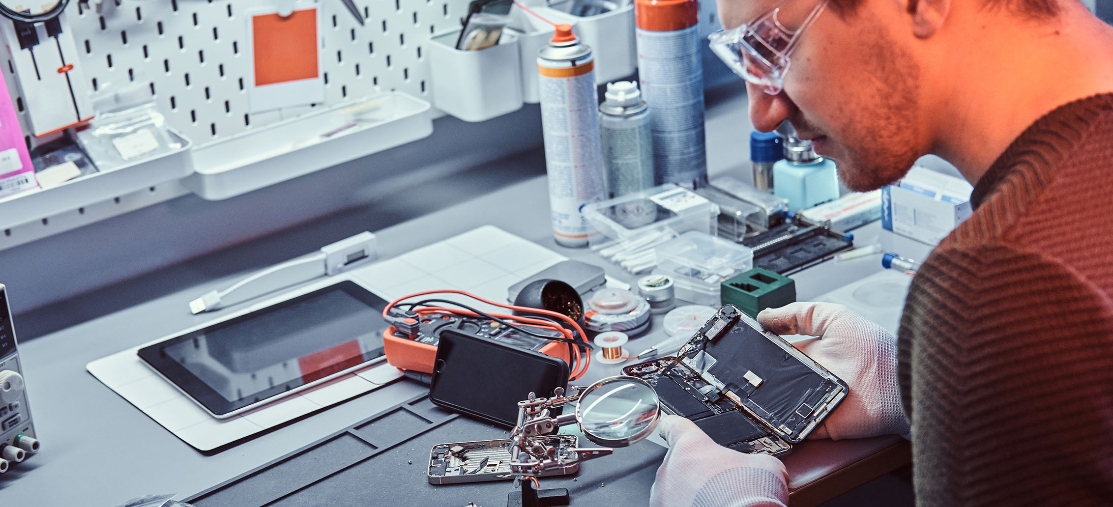Technicien portant des gants et des lunettes de protection, réparant un smartphone ouvert sur un bureau équipé d'outils électroniques et de matériel de réparation.