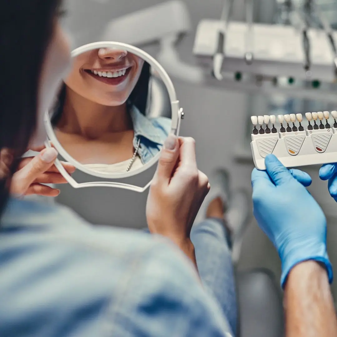 A dentist holds a shade guide while showing a patient their reflection in a handheld mirror inside a dental clinic.