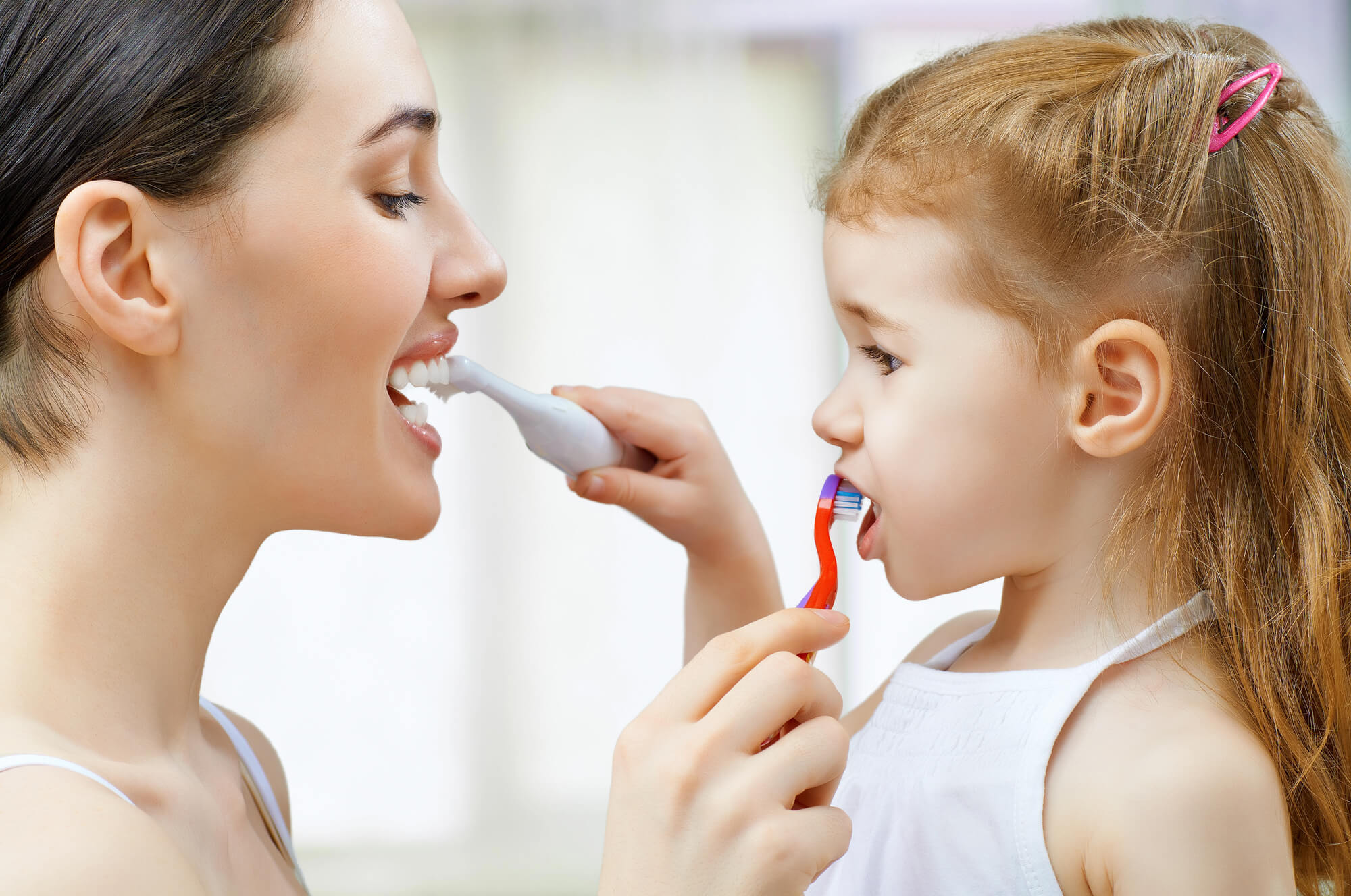 A child playfully applies cream to the arm of an adult, both wearing white tops in a softly lit room.