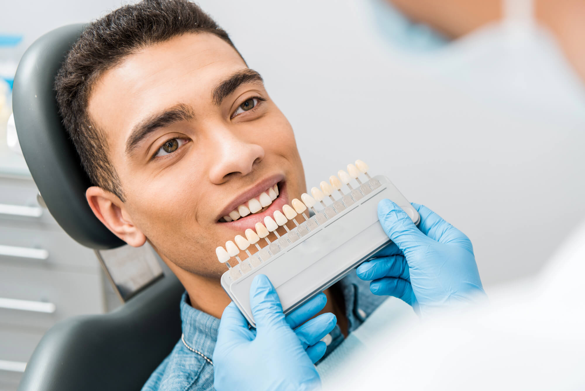 A dental professional in blue gloves holds a shade guide, comparing colors with a patient seated in a dental chair.
