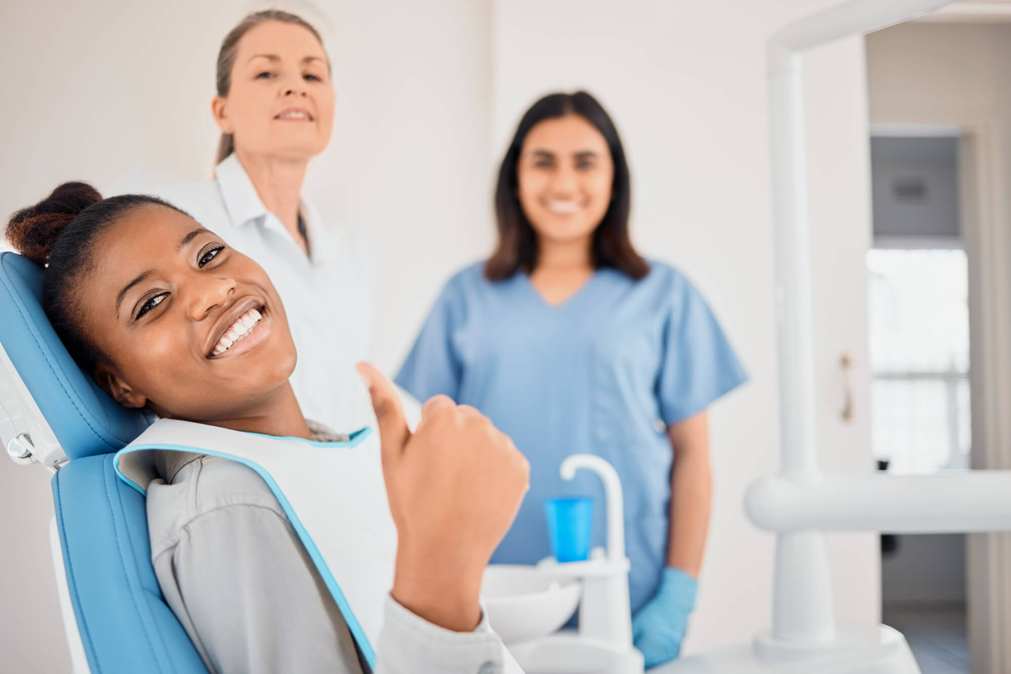 A patient in a dental chair gives a thumbs up, while a dentist and dental assistant stand nearby, ready for the appointment.