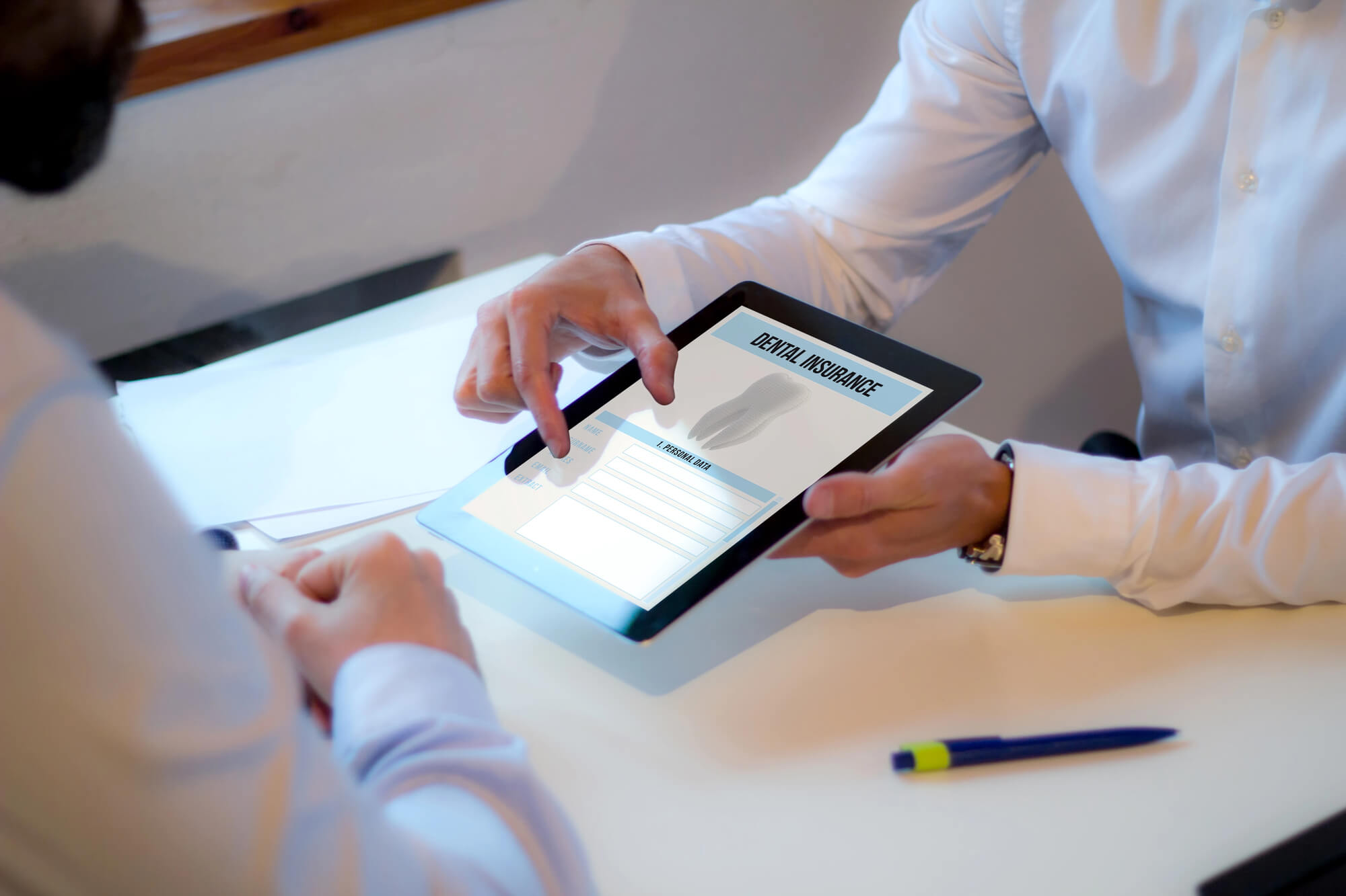 Two people discussing dental insurance on a tablet during a meeting at a desk, with paperwork and a pen visible.