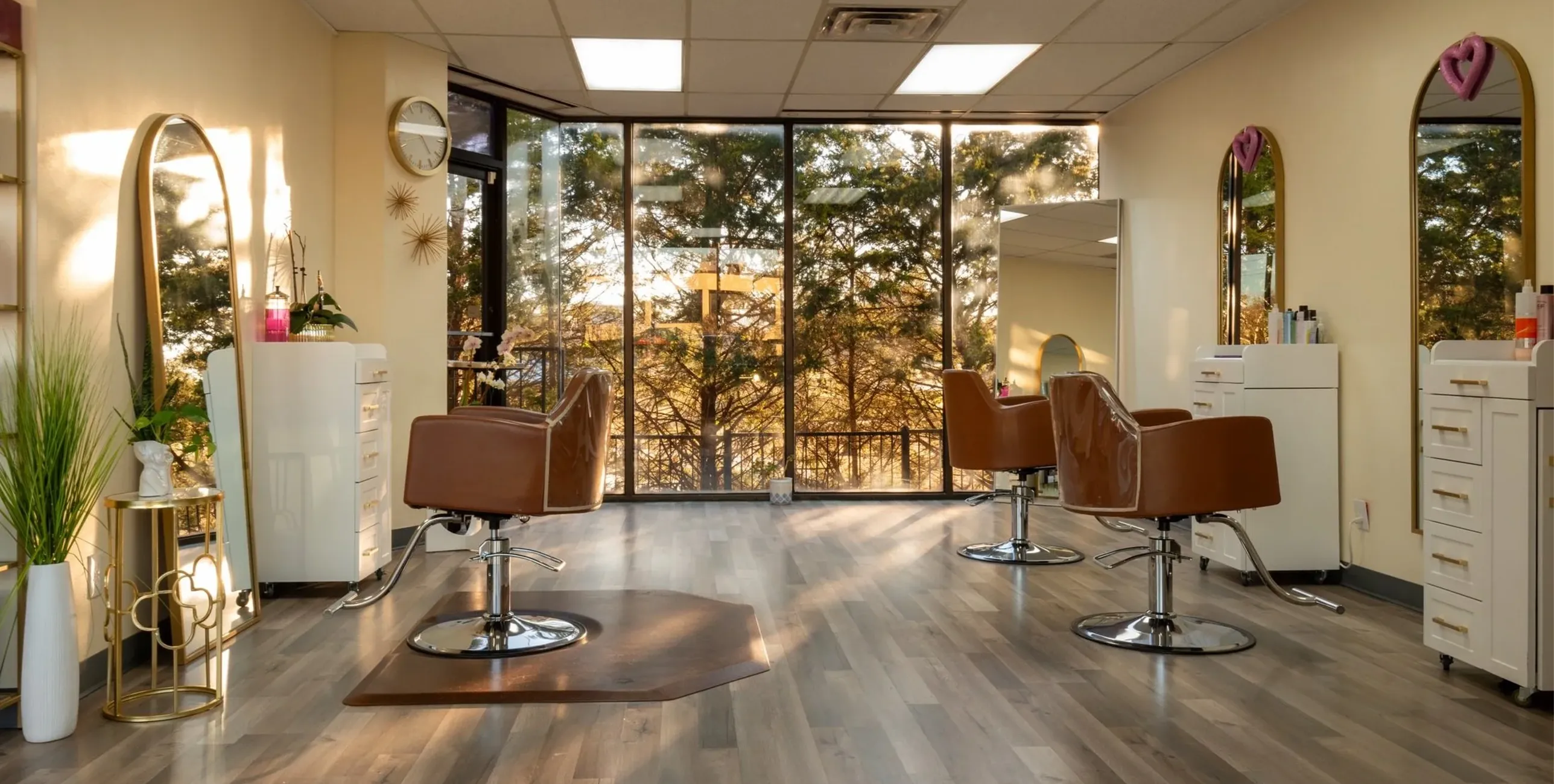 Bright hair salon with large windows, three brown styling chairs, mirrors, and white cabinets.