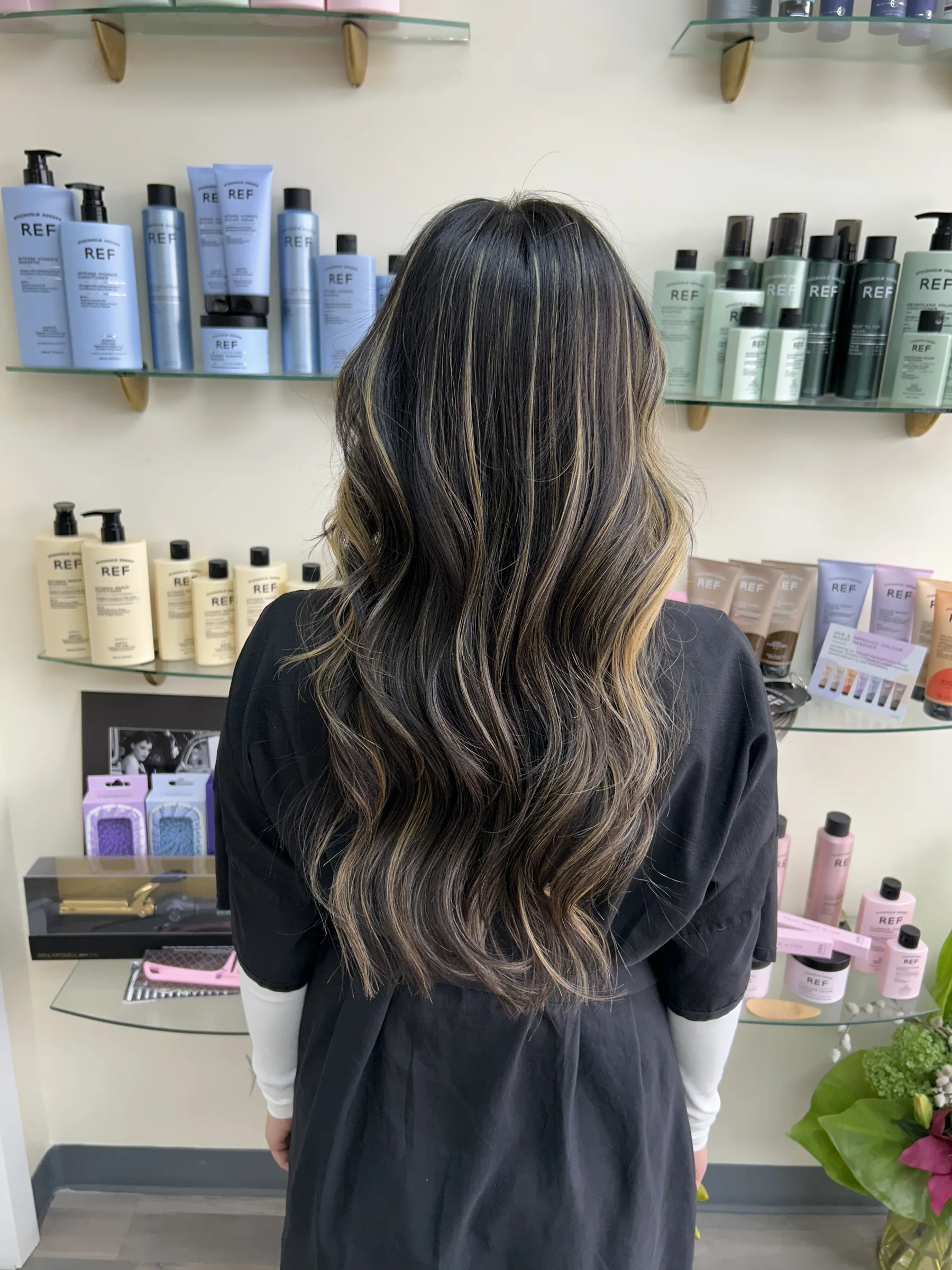 Person with long, wavy dark hair with blonde highlights standing in front of shelves with haircare products.