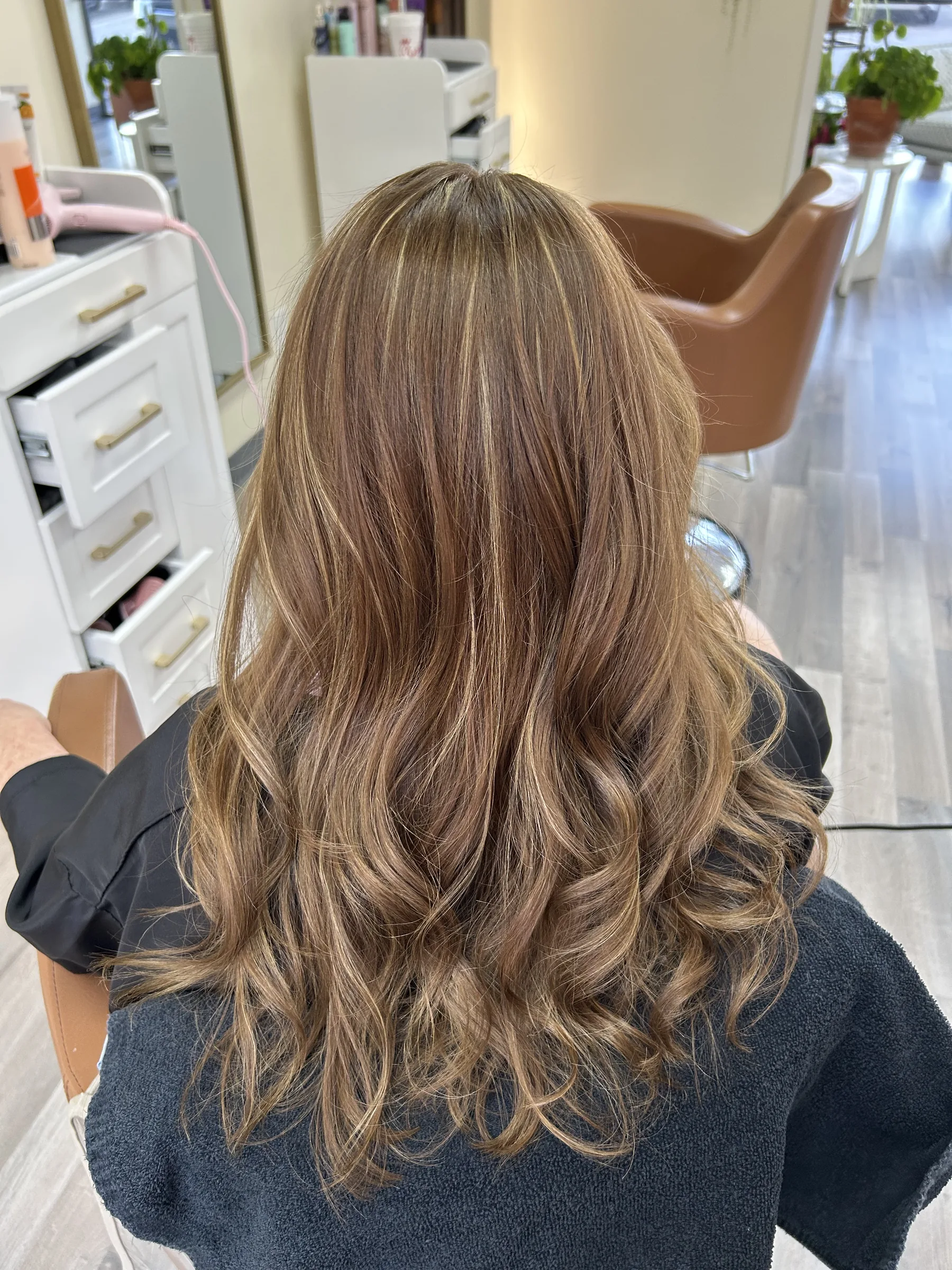 Back view of a person with long, wavy, highlighted brown hair sitting in a salon chair.