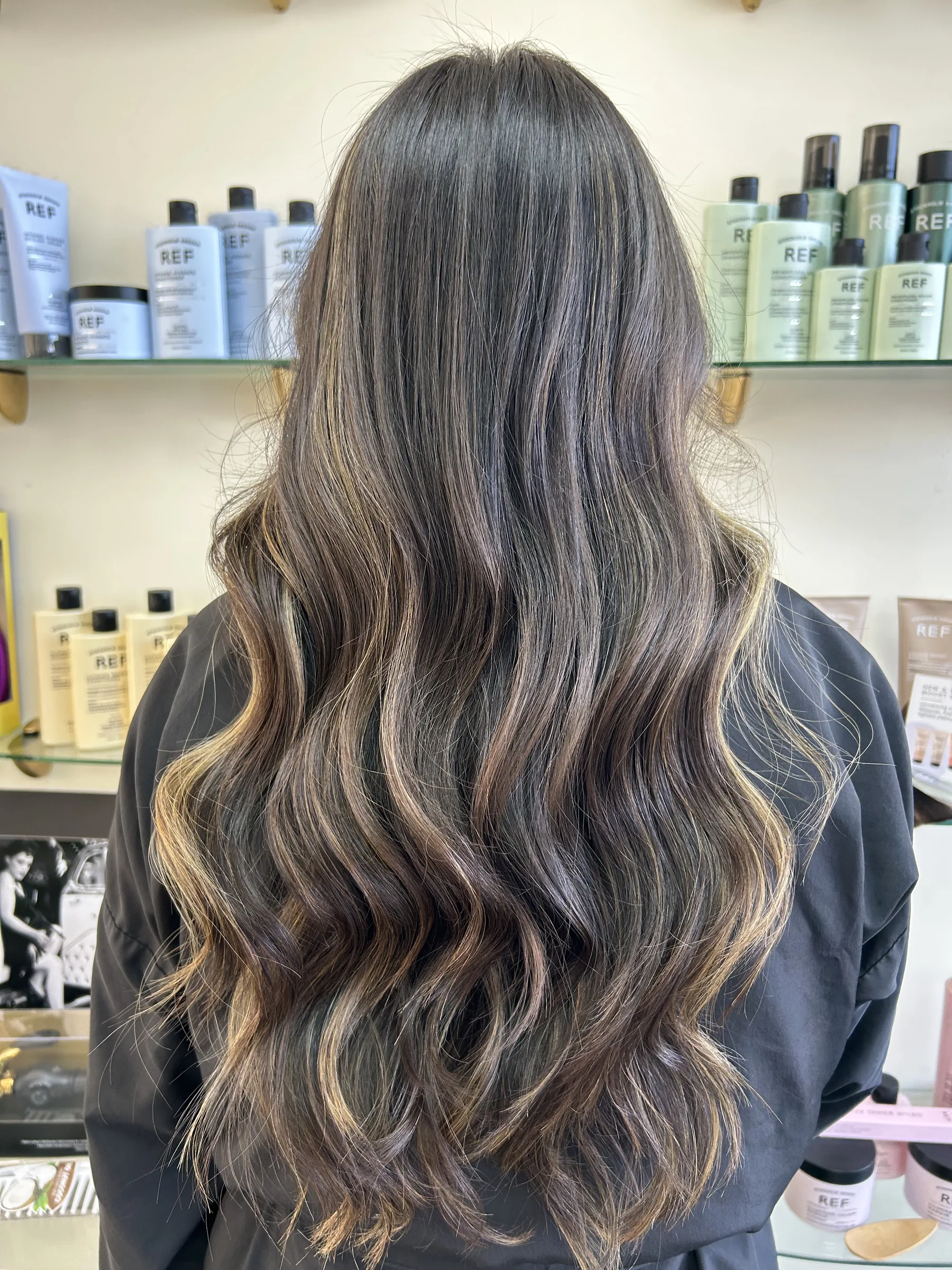 Long wavy brown hair with blonde highlights on a person wearing a black cape in a salon.