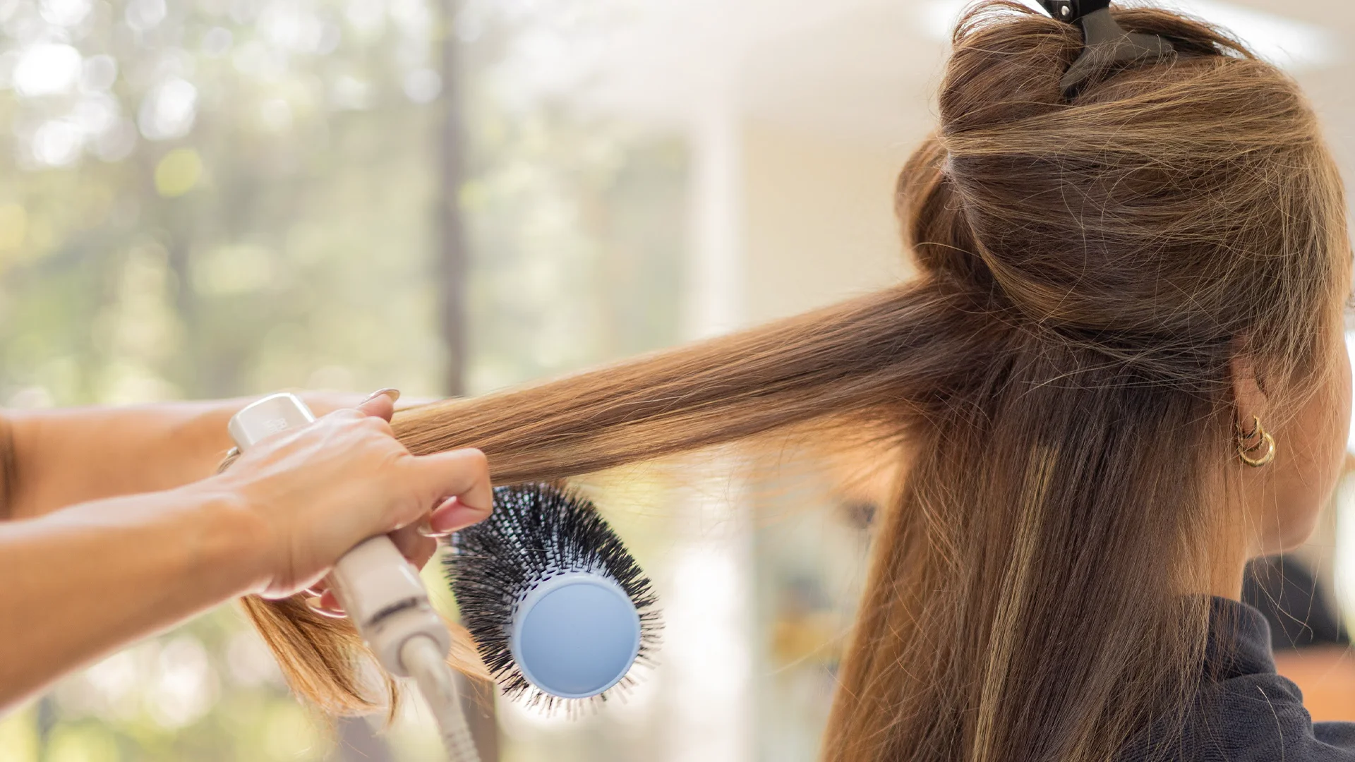 Hands blow drying and brushing long brown hair section in a bright salon.